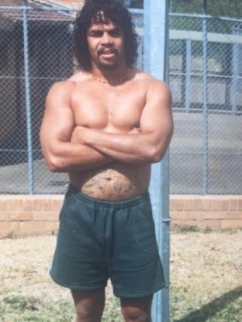 A man wearing shorts and no shirt stands with arms folded in jail